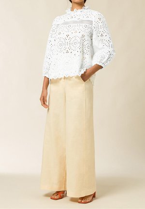 Femme portant un chemisier blanc en dentelle à œillets, un pantalon large beige et des sandales à talons orange, une main dans la poche.