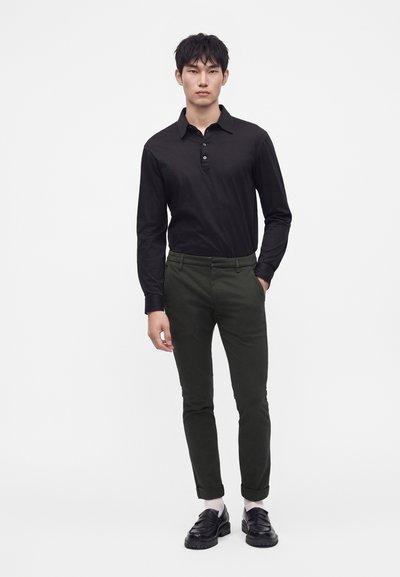 Jeune homme debout portant un polo noir à manches longues, un pantalon slim vert foncé, des chaussettes blanches et des mocassins noirs sur fond blanc.
