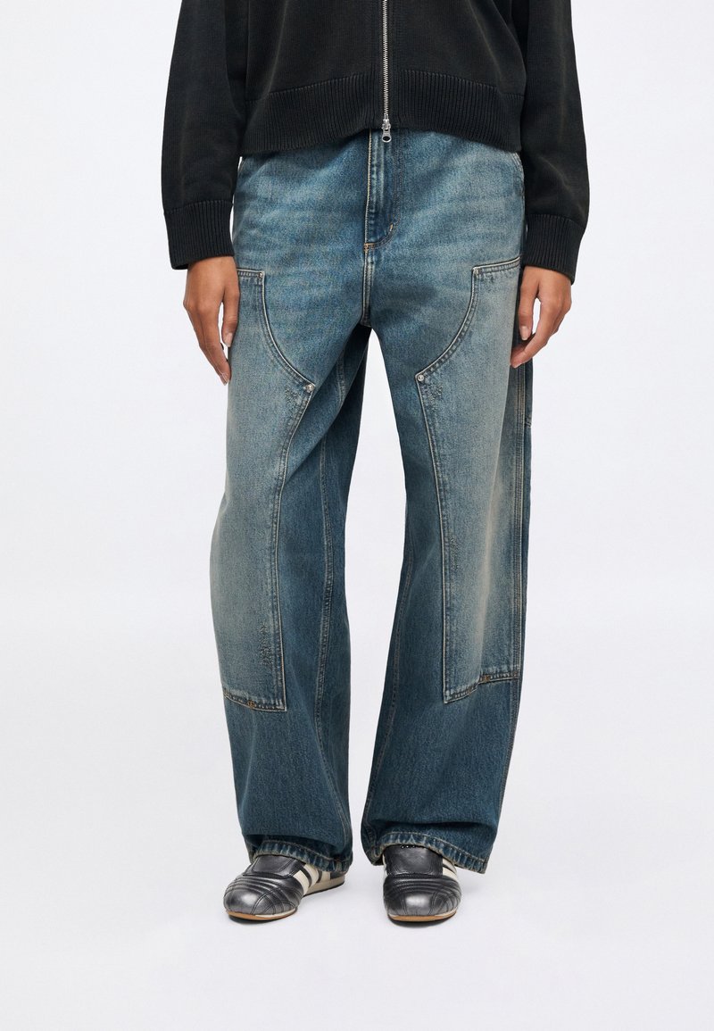 Løse blå denimjeans med let falming, der har markante buede sømme og lommer. Kombineret med grå sko og en sort lynlåstrøje.