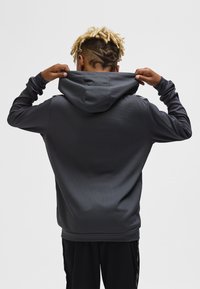 Person mit blonden Dreadlocks und rasiertem, gemustertem Undercut, der einen dunkelgrauen Kapuzenpullover trägt und die Kapuze von hinten hochhält.