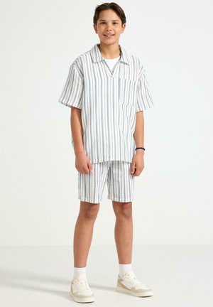 Poika seisoo, päällään valko-harmaaraidallinen lyhythihainen paita ja siihen sopivat shortsit, valkoiset sukat ja beigejä lenkkareita.