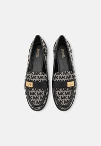 Une paire de mocassins noirs et beiges de Michael Kors avec un motif logo MK et une quincaillerie en ton doré sur un fond gris.