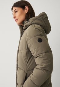 Frau, die nach links schaut, trägt eine olivgrüne gepolsterte Winterjacke mit Kapuze, die Kordeln hat, und ein schwarzes rundes Logo auf dem Ärmel.