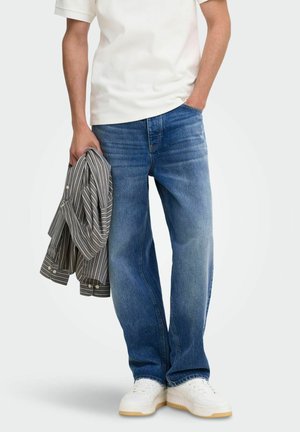 Personne portant des baskets blanches, un jean bleu ample et une chemise blanche, tenant dans une main une chemise rayée à boutons pliée.