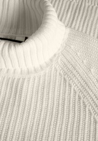 Baltas megztinis su raukšlėta tekstūra, aukštu apykakle ir įstrižais raštų akcentais. Medžiaga atrodo minkšta ir stora.