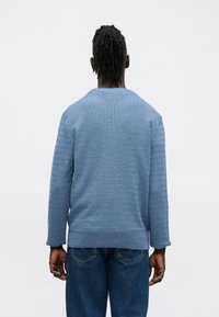 Zils teksturēts džemperis ar garām piedurknēm, apaļu kakla izgriezumu un brīvu piegriezumu, kombinēts ar tumši zilām džinsa biksēm. Skats no aizmugures izceļ adījuma rakstu.