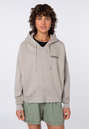 Junge Person mit lockigem Haar trägt einen hellgrauen Zip-Hoodie mit dem Schriftzug „Love & Passion“ und olivgrünen Shorts, steht vor weißem Hintergrund.