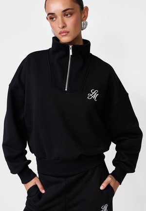 Žena oblečená v čiernej mikine na zips a zodpovedajúcich nohaviciach s bielym logom „LM“, ruky v vreckách a veľké strieborné kruhové náušnice.