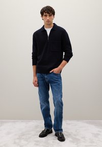 Pullover navy con colletto a zip e tasca sul petto, abbinato a jeans azzurri a gamba dritta e mocassini neri su uno sfondo grigio chiaro.