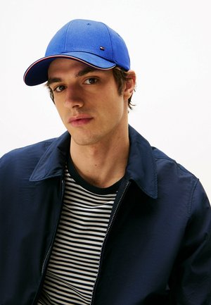 Blaues Baseballcap mit gebogenem Schirm und roten Akzenten, getragen mit einer marineblauen Jacke und gestreiftem Hemd. Glatter Stoff mit lässiger Passform.