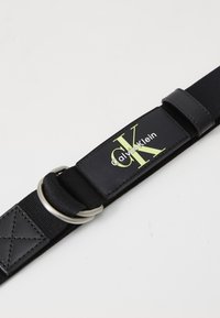 Black elastic strap with a rectangular leather patch and silver metal ring; features "Calvin Klein" logo in green and white text.