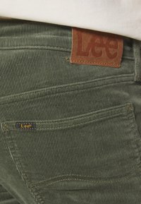 Pantalones de pana verdes con una etiqueta de cuero marrón con "Lee" grabado. Incluyen una pequeña etiqueta negra con texto amarillo en un bolsillo.
