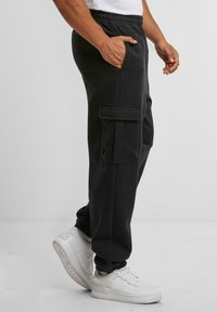 Black cargo sweatpants with elastic waistband, side pockets, and a flap pocket. Fabric has a smooth texture. Paired with white sneakers.