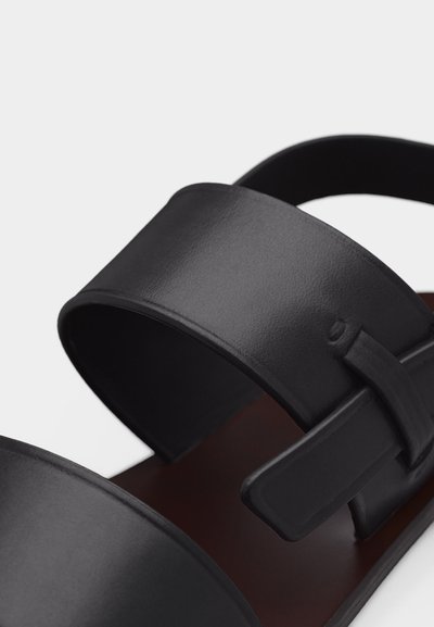Κοντινό πλάνο μαύρου δερμάτινου σανδαλιού με φαρδιές λουριά και λεπτομέρεια με κλειδί σε λευκό φόντο.