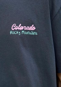 Grå T-shirt med korte ærmer; har "Colorado" broderet i pink og "Rocky Mountains" broderet i teal på brystområdet.
