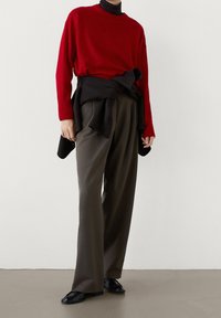 Punainen villaneule mustan poolopaidan päällä, yhdistettynä löysiin oliivinvihreisiin housuihin ja mustiin kenkiin. Yksinkertainen, moderni muotoilu.