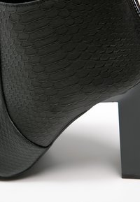 Gros plan d'une chaussure à talon haut noire avec un motif texturé rappelant la peau de reptile et un talon carré solide sur une surface blanche.