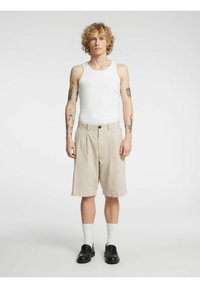 Beige shorts med folder og svart knappelukking, kombinert med en hvit tanktopp, svarte loafers og hvite ankelstrømper. Glatt tekstur.