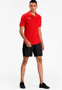 Rød polo skjorte med krave og korte ærmer, med et hvidt logo. Sættes sammen med sorte shorts og sorte sportsko.