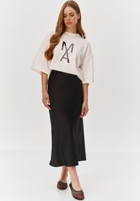 Biała, oversize koszulka z czarnym graficznym wzorem i luźnymi rękawami, zestawiona z czarną satynową spódnicą do kostek i czarnymi płaskimi butami z siatki.
