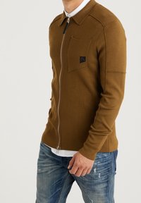 Hnědý pletený cardigan na zip s límcem, obsahující náprsní kapsu a logo. Nosený přes bílou košili, s texturovanou tkaninou.