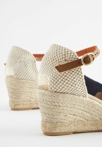 Sandalias de cuña con parte superior de tejido trenzado en beige y blanco y correa de tobillo de cuero marrón. La plataforma cubierta de yute añade textura y altura.