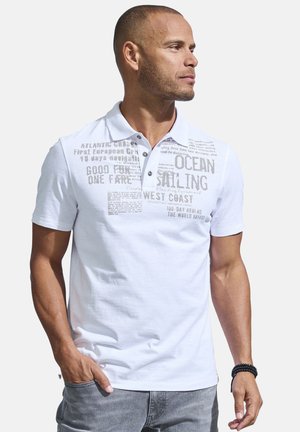 Mann trägt weißes Poloshirt mit Segelmotiv-Schriftzug, schaut nach rechts, Hand in der Tasche, vor einfachem Hintergrund.