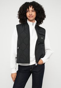 Juodas siuvinėtas liemenė su stovinčia apykakle, užtrauktuku ir šoninėmis kišenėmis, dėvima virš balto užtrauktuko megztinio, derinama su tamsiomis džinsinėmis kelnėmis.