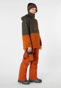 Schneejacke mit grauen und orangefarbenen Einsätzen, Reißverschlusstaschen, elastischen Bündchen. Kombiniert mit orangenen Hosen und grauen Stiefeln, hält schwarze Fäustlinge in der Hand.