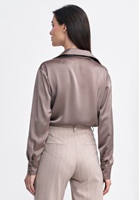 Satynowa bluzka w ciemnym taupe z delikatnym połyskiem, z długimi rękawami i klasycznym kołnierzykiem, w zestawie z jasnobeżowymi spodniami w prążki.