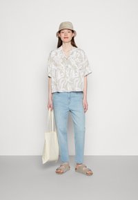 Camisa de manga curta com padrão floral claro, jeans azul claro, chapéu estilo balde bege, sandálias bege e uma bolsa de tela bege.