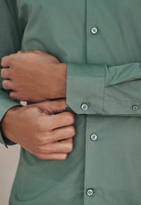 Grünes Langarmhemd aus glattem Stoff mit klassischem Kragen, Knopfleiste und dezenten Nähten sowie Knopfakzenten an den Manschetten.