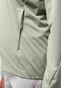 Veste vert clair au toucher lisse, avec une poche zippée sur le côté et une coupe ajustée. Le tissu semble léger et flexible.
