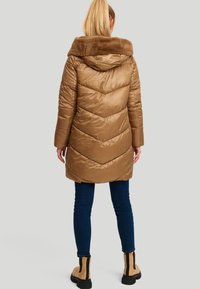 Vrouw met een blonde paardenstaart, gekleed in een bruine gewatteerde jas met een faux fur kraag, donkere spijkerbroek en tan laarzen met zwarte zolen, staand met de rug naar de kijker.