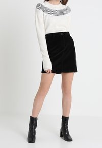 Svart kordflugekjol med knäppning framtill. Matchas med en vit stickad tröja med en svart geometrisk design vid halsringningen.