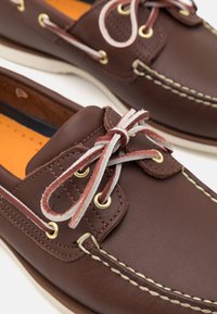 Braune Lederschuhe für Boote mit strukturierten Oberflächen, goldenen Ösen und kontrastierenden grauen Schnürsenkeln. Orangene Einlegesohlen sind sichtbar. Traditionelles Design.
