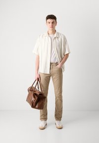 Beige katoenen broek, wit T-shirt, gestreept kortärmelig overhemd en bruine leren handtas. Model draagt witte sneakers met beige accenten.