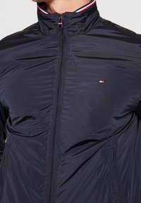 Marineblauwe ritsjack gemaakt van lichtgewicht materiaal, met een hoge kraag met rode en witte strepen, en een klein embleem op de borst.