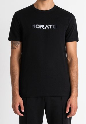 Uomo che indossa una maglietta nera con il logo "MORATO" in lettere lucide e scure centrate sul petto su uno sfondo bianco semplice.