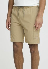 Beige cargo shorts with elastic waistband and drawstring, featuring side pockets and a patch pocket on the thigh with logo detail.