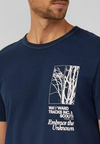 Muškarac nosi majicu tamnoplave boje s bijelim grafičkim prikazom grančica drveta i natpisom "WAYWARD TRACKS INC. & SCOUTS Prihvatite nepoznato" na prsima.