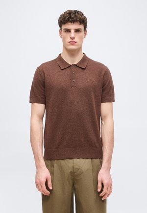 Jeune homme debout, droit, portant un polo marron à manches courtes et un pantalon vert olive, sur un fond clair uni.