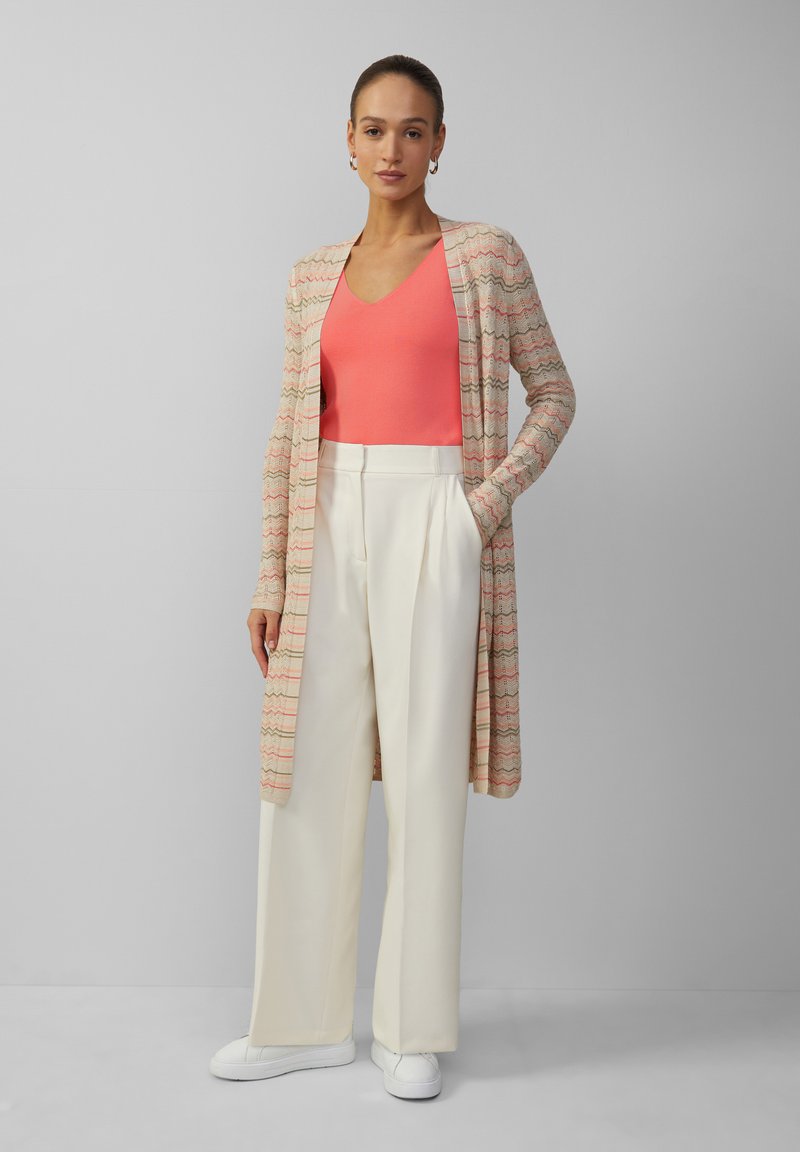 Langer, offener Cardigan in Creme mit buntem Zickzackmuster, kombiniert mit einem korallenfarbenen Oberteil und weißen, weiten Hosen, abgerundet mit weißen Sneakers.