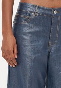 Dark denim jeans with yellow stitching, straight-leg design, front pockets, and a silver button closure. Fabric has a slight sheen.