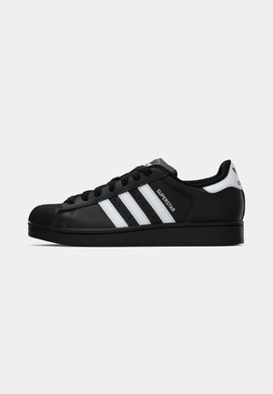 Black Adidas Superstar sneaker with white stripes, shell toe, and "Superstar" text on side, shown in side profile on white background.