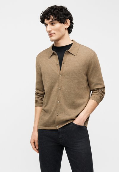 Νέος άνδρας με σγουρά μαλλιά που φοράει ένα ταμπά μπουφάν με κουμπιά πάνω από ένα μαύρο πουκάμισο και σκούρα τζιν, με το χέρι στην τσέπη, κοιτάζοντας στο πλάι.