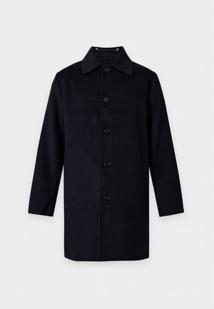 Un homme porte un manteau noir sur un cardigan gris et une chemise rouge, avec un pantalon foncé et des mocassins noirs, sur un fond blanc uni.