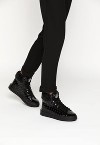 Černé vysoké tenisky s lesklým lakovaným povrchem, prošívanou texturou, kožešinovým límcem a černou gumovou podrážkou. Nohou s černými kalhotami.