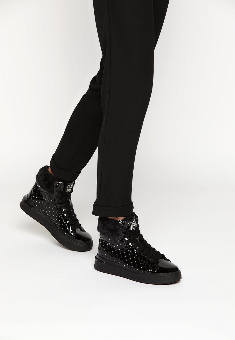 Černé vysoké tenisky s lesklým lakovaným povrchem, prošívanou texturou, kožešinovým límcem a černou gumovou podrážkou. Nohou s černými kalhotami.