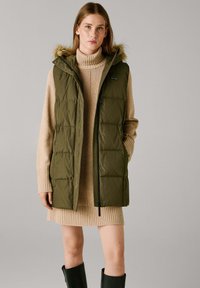 Oliwkowa kamizelka puchowa z kapturem, ozdobiona futrzanym wykończeniem i zapięciem na zamek, noszona na beżowej sukience swetrowej w prążki oraz czarnych kozakach za kolano.